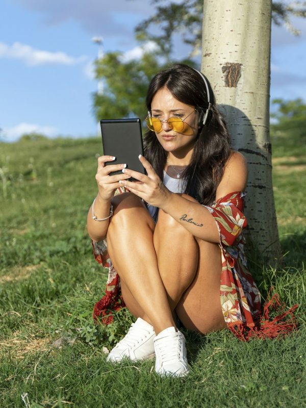 Woman with sunglasses and headphones reading an ebook while relaxing sitting on grass in a park